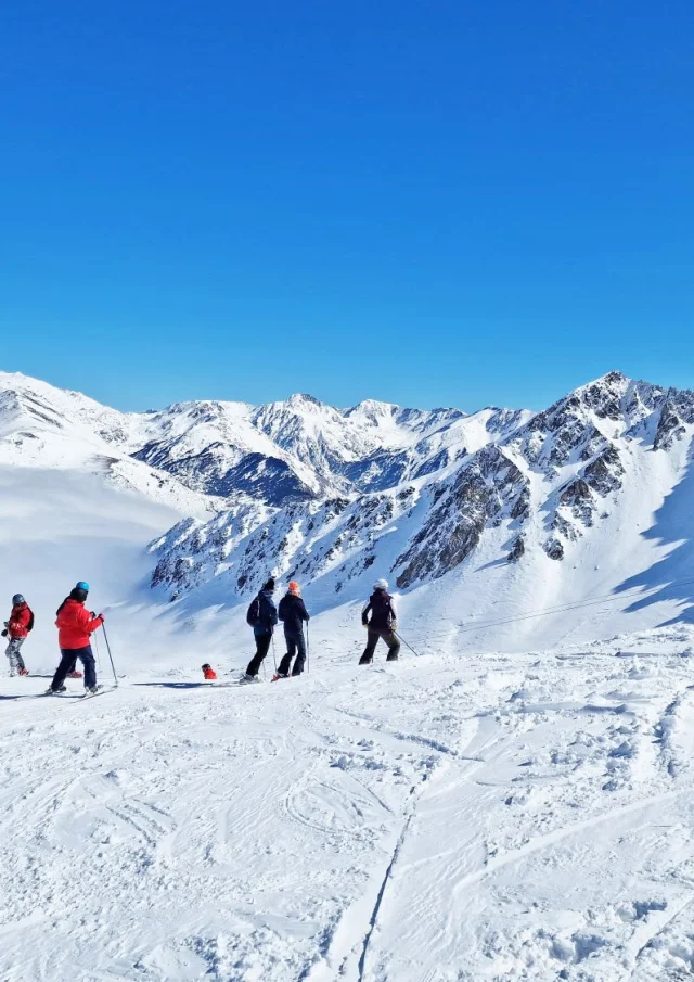 Groupe de skieurs sur une piste enneigée dominant une mer de nuages, entourée de hauts sommets sous un ciel bleu.