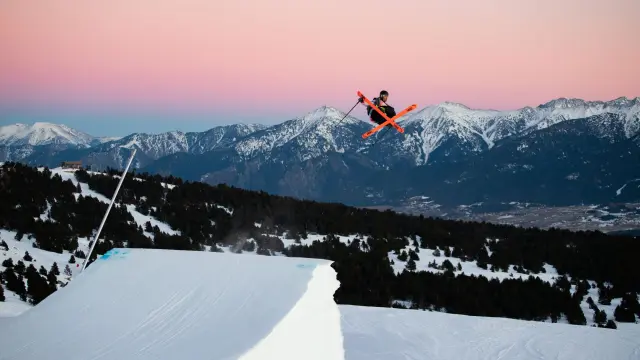 Skieur effectuant une figure aérienne au snowpark de Font-Romeu, avec en arrière-plan les montagnes enneigées au coucher du soleil.