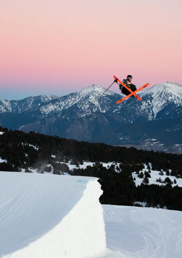 Skieur effectuant une figure aérienne au snowpark de Font-Romeu, avec en arrière-plan les montagnes enneigées au coucher du soleil.