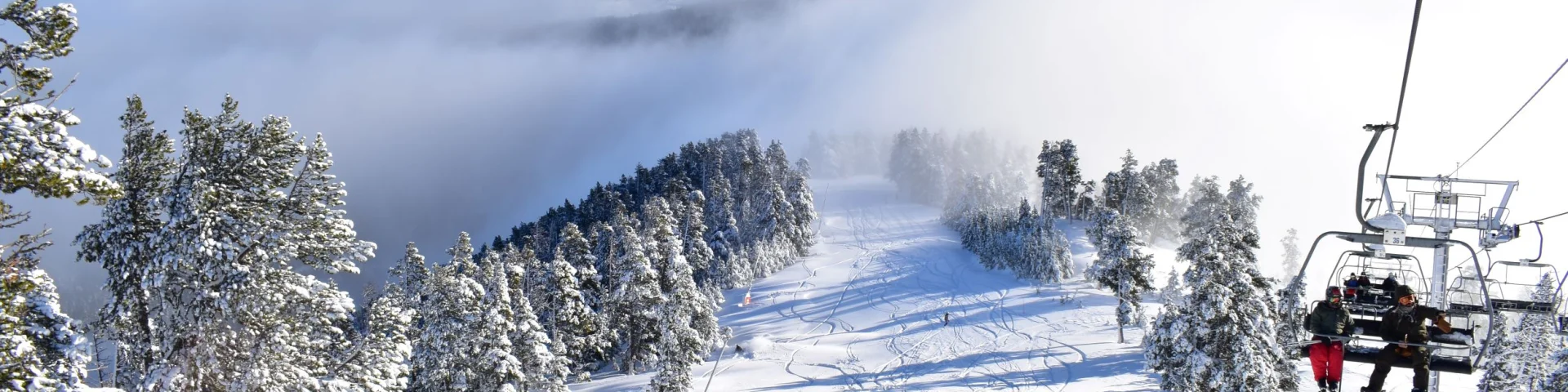 Skieurs sur une piste enneigée bordée d’arbres, avec un télésiège en fonctionnement et une mer de nuages recouvrant les vallées en contrebas sous un ciel bleu.