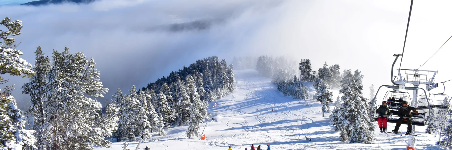 Skieurs sur une piste enneigée bordée d’arbres, avec un télésiège en fonctionnement et une mer de nuages recouvrant les vallées en contrebas sous un ciel bleu.
