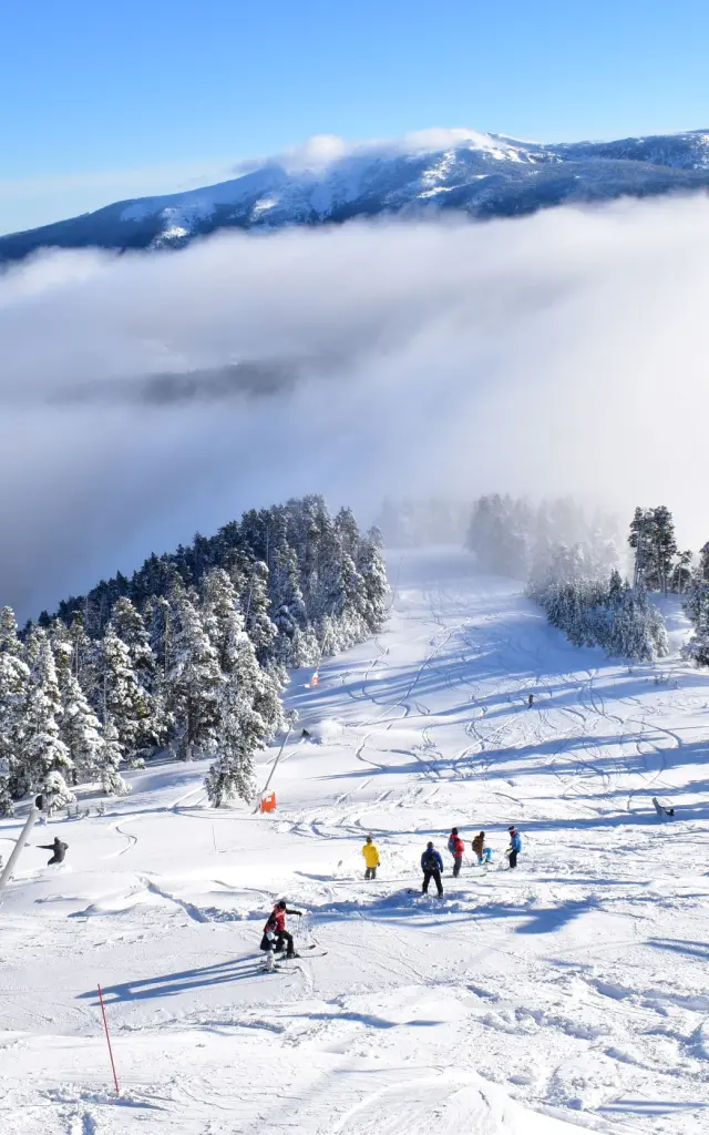 Skieurs sur une piste enneigée bordée d’arbres, avec un télésiège en fonctionnement et une mer de nuages recouvrant les vallées en contrebas sous un ciel bleu.