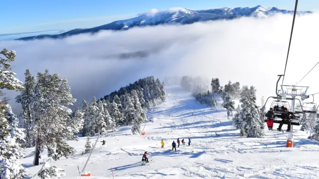 Skieurs sur une piste enneigée bordée d’arbres, avec un télésiège en fonctionnement et une mer de nuages recouvrant les vallées en contrebas sous un ciel bleu.