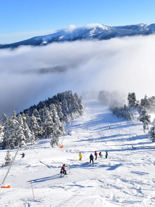 Skieurs sur une piste enneigée bordée d’arbres, avec un télésiège en fonctionnement et une mer de nuages recouvrant les vallées en contrebas sous un ciel bleu.