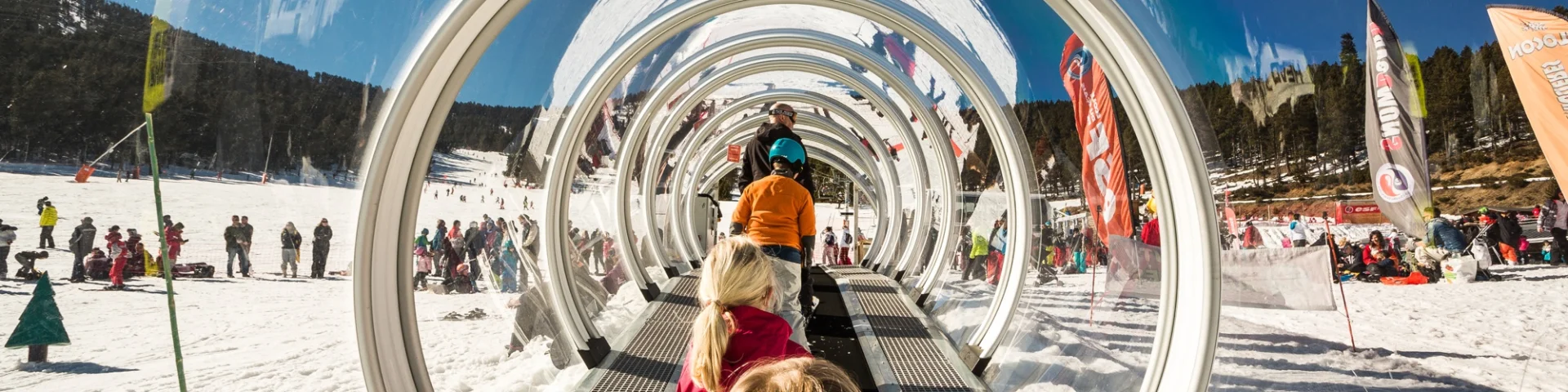 Des enfants montent sur un tapis roulant couvert transparent dans une station de ski enneigée, entourés d’autres skieurs et de familles sous un ciel bleu.