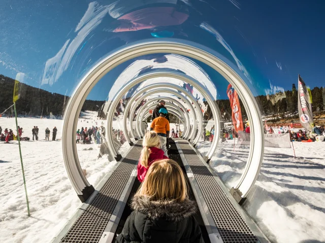 Des enfants montent sur un tapis roulant couvert transparent dans une station de ski enneigée, entourés d’autres skieurs et de familles sous un ciel bleu.