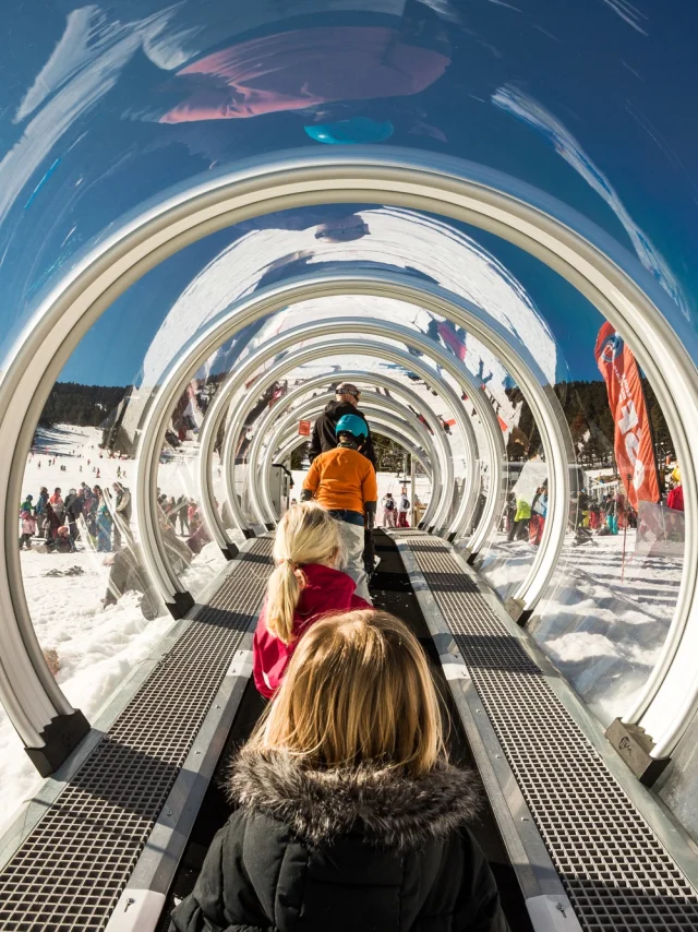 Des enfants montent sur un tapis roulant couvert transparent dans une station de ski enneigée, entourés d’autres skieurs et de familles sous un ciel bleu.