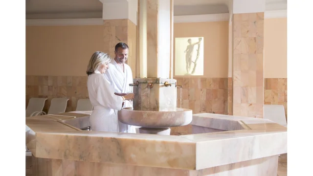 Un couple en peignoir se sert de l’eau thermale à une fontaine intérieure en marbre, dans un espace de repos lumineux des Thermes de La Preste.