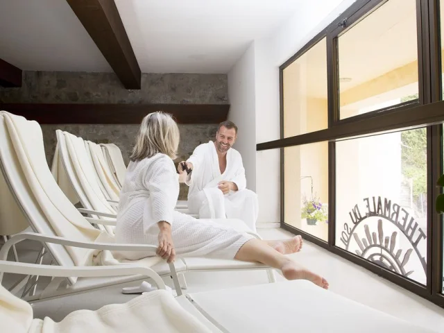 Deux curistes en peignoir blanc se détendent sur des transats à l’intérieur des Thermes de La Preste, profitant d’un moment de repos face à une grande baie vitrée.