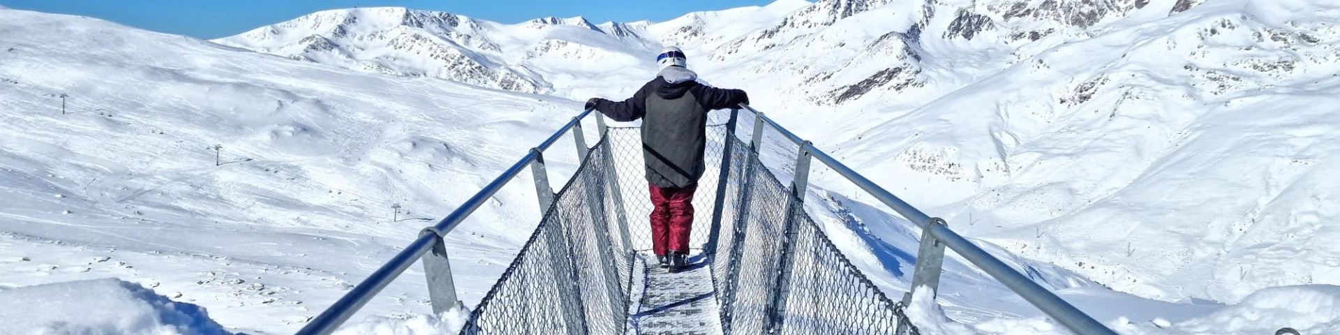 Personne debout sur la passerelle panoramique Le Pied dans le Vide à Porté-Puymorens, face aux montagnes enneigées sous un ciel bleu.