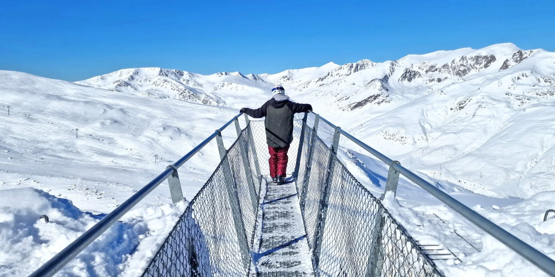 Personne debout sur la passerelle panoramique Le Pied dans le Vide à Porté-Puymorens, face aux montagnes enneigées sous un ciel bleu.