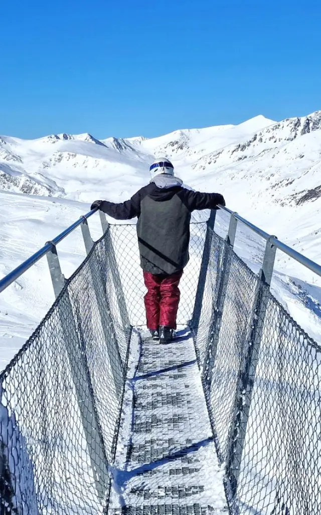 Personne debout sur la passerelle panoramique Le Pied dans le Vide à Porté-Puymorens, face aux montagnes enneigées sous un ciel bleu.