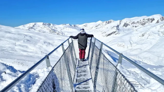 Personne debout sur la passerelle panoramique Le Pied dans le Vide à Porté-Puymorens, face aux montagnes enneigées sous un ciel bleu.