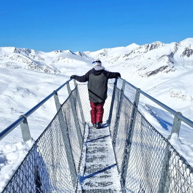 Personne debout sur la passerelle panoramique Le Pied dans le Vide à Porté-Puymorens, face aux montagnes enneigées sous un ciel bleu.