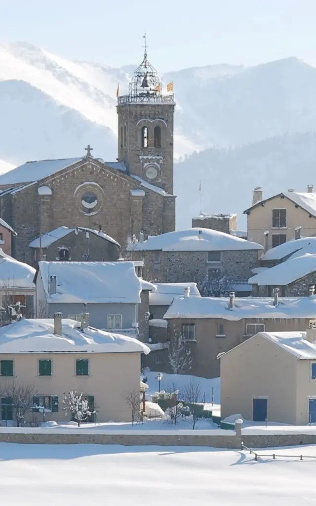 Vue sur le village de Font-Romeu couvert de neige, avec son église et les montagnes enneigées en arrière-plan.