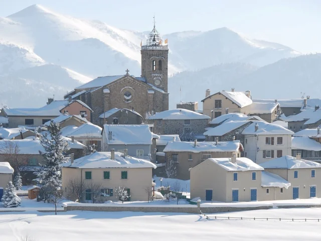 Vue sur le village de Font-Romeu couvert de neige, avec son église et les montagnes enneigées en arrière-plan.