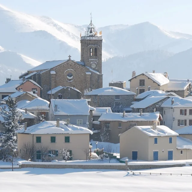 Vue sur le village de Font-Romeu couvert de neige, avec son église et les montagnes enneigées en arrière-plan.