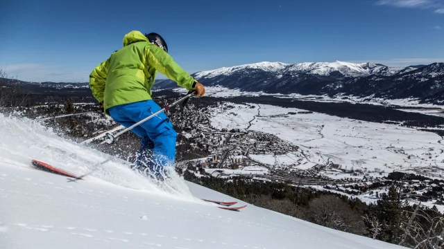 Skieur en descente sur une piste enneigée surplombant le village des Angles et la vallée recouverte de neige, sous un ciel bleu.
