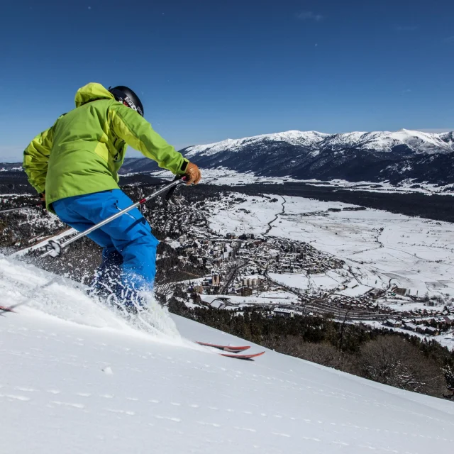 Skieur en descente sur une piste enneigée surplombant le village des Angles et la vallée recouverte de neige, sous un ciel bleu.