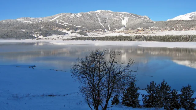 Vue sur le lac de Matemale partiellement gelé, avec le village et les pistes de la station de ski des Angles en arrière-plan sous un ciel bleu.