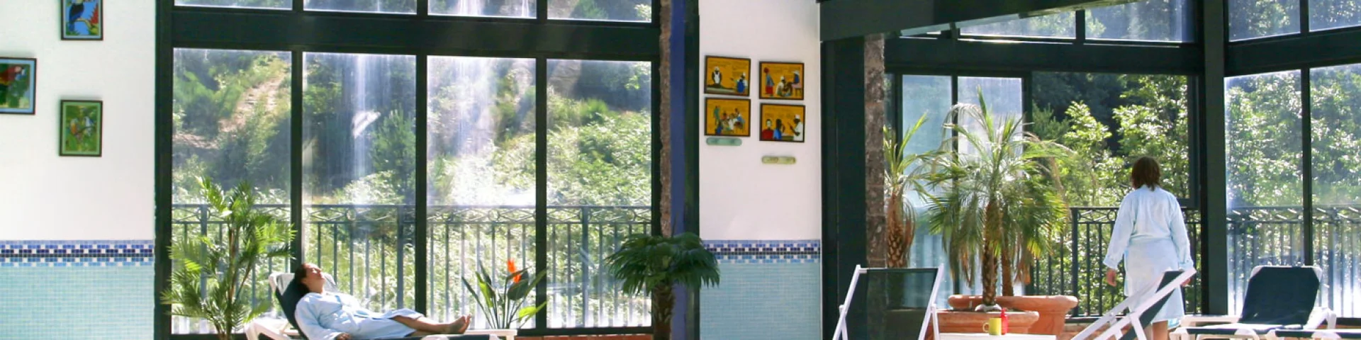 Espace bien-être intérieur du Domaine de Falgos, avec piscine chauffée, baies vitrées ouvertes sur la nature et une cascade visible à l’extérieur. Deux personnes se détendent en peignoir au bord de l’eau.