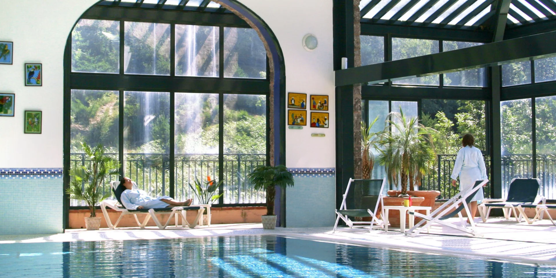 Espace bien-être intérieur du Domaine de Falgos, avec piscine chauffée, baies vitrées ouvertes sur la nature et une cascade visible à l’extérieur. Deux personnes se détendent en peignoir au bord de l’eau.