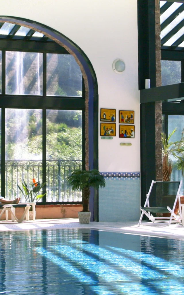 Espace bien-être intérieur du Domaine de Falgos, avec piscine chauffée, baies vitrées ouvertes sur la nature et une cascade visible à l’extérieur. Deux personnes se détendent en peignoir au bord de l’eau.