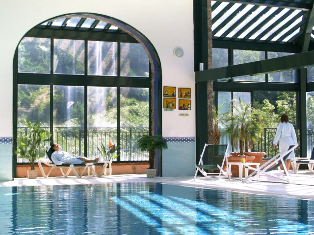 Espace bien-être intérieur du Domaine de Falgos, avec piscine chauffée, baies vitrées ouvertes sur la nature et une cascade visible à l’extérieur. Deux personnes se détendent en peignoir au bord de l’eau.