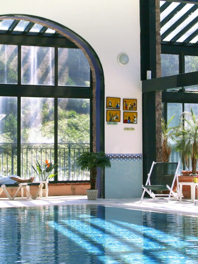 Espace bien-être intérieur du Domaine de Falgos, avec piscine chauffée, baies vitrées ouvertes sur la nature et une cascade visible à l’extérieur. Deux personnes se détendent en peignoir au bord de l’eau.