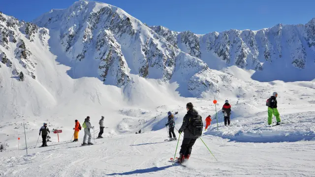 Skieurs sur une piste enneigée au pied des sommets escarpés de la station de Porté-Puymorens, sous un ciel bleu dégagé.