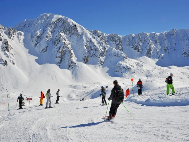 Skieurs sur une piste enneigée au pied des sommets escarpés de la station de Porté-Puymorens, sous un ciel bleu dégagé.