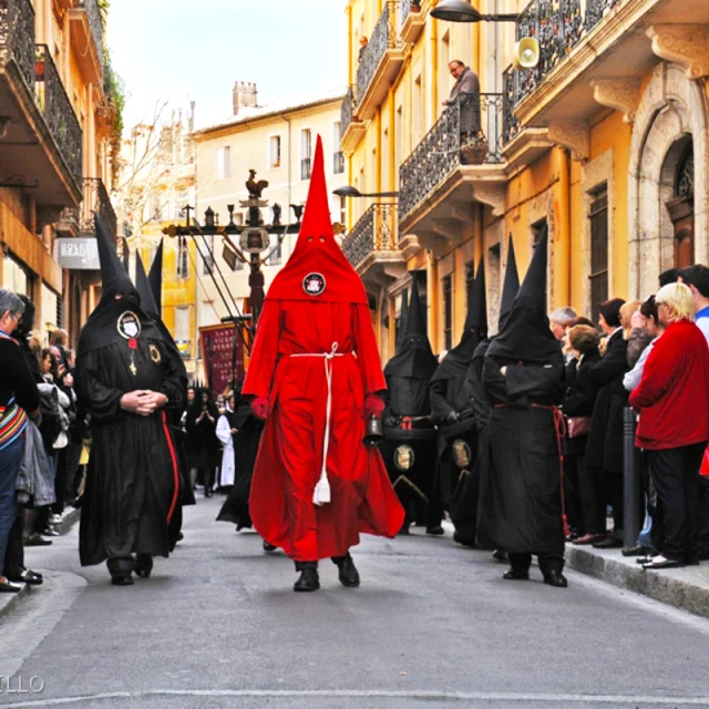 Défilé de pénitents vêtus de capuchons pointus noirs et rouges lors de la Procession de la Sanch à Perpignan, dans une rue bordée de bâtiments anciens et de spectateurs.