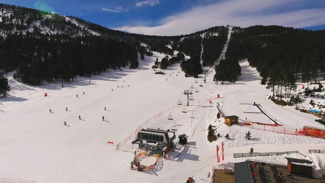 Vue aérienne d’une station de ski entourée de forêts de pins, avec plusieurs skieurs sur les pistes enneigées et un télésiège en fonctionnement sous un ciel bleu.