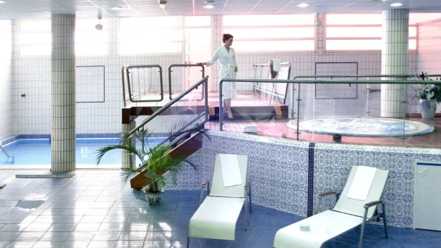 Espace bien-être intérieur des Thermes de Vernet-les-Bains avec piscine, jacuzzi, transats et une femme en peignoir se dirigeant vers les bassins.