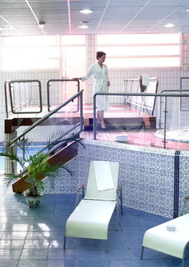 Espace bien-être intérieur des Thermes de Vernet-les-Bains avec piscine, jacuzzi, transats et une femme en peignoir se dirigeant vers les bassins.