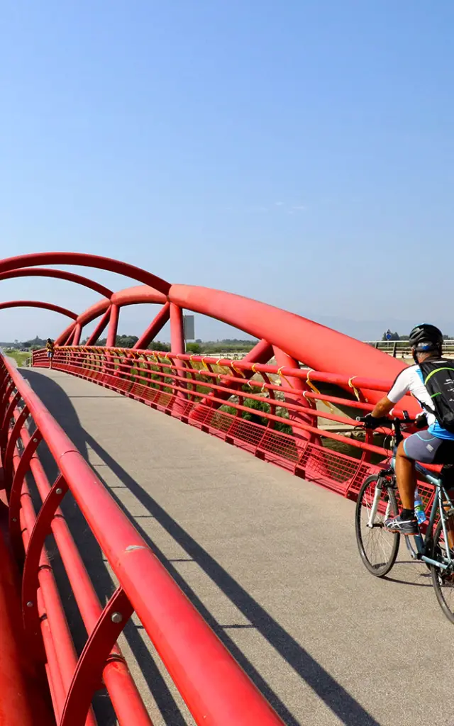 Cycliste traversant une passerelle rouge au-dessus d’un cours d’eau, dans un paysage ouvert.