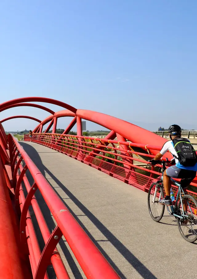 Cycliste traversant une passerelle rouge au-dessus d’un cours d’eau, dans un paysage ouvert.