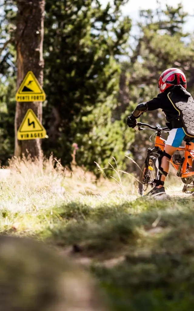 Enfant équipé de protections descendant à VTT sur un sentier, avec panneaux signalant pente forte et virages.