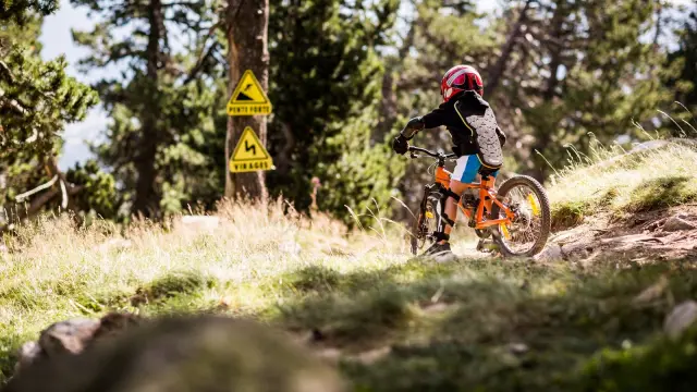 Enfant équipé de protections descendant à VTT sur un sentier, avec panneaux signalant pente forte et virages.