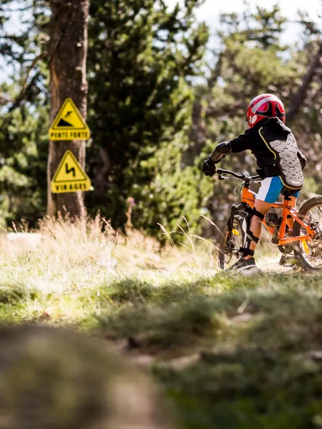 Enfant équipé de protections descendant à VTT sur un sentier, avec panneaux signalant pente forte et virages.