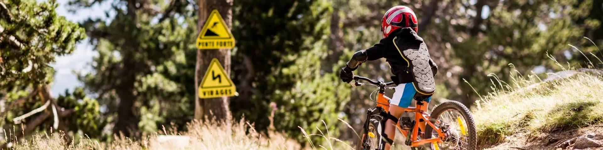 Enfant équipé de protections descendant à VTT sur un sentier, avec panneaux signalant pente forte et virages.