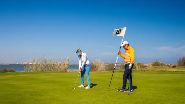 Deux golfeurs sur un green en bord d’étang, sous un ciel bleu dégagé, l’un prêt à putter, l’autre tenant le drapeau.