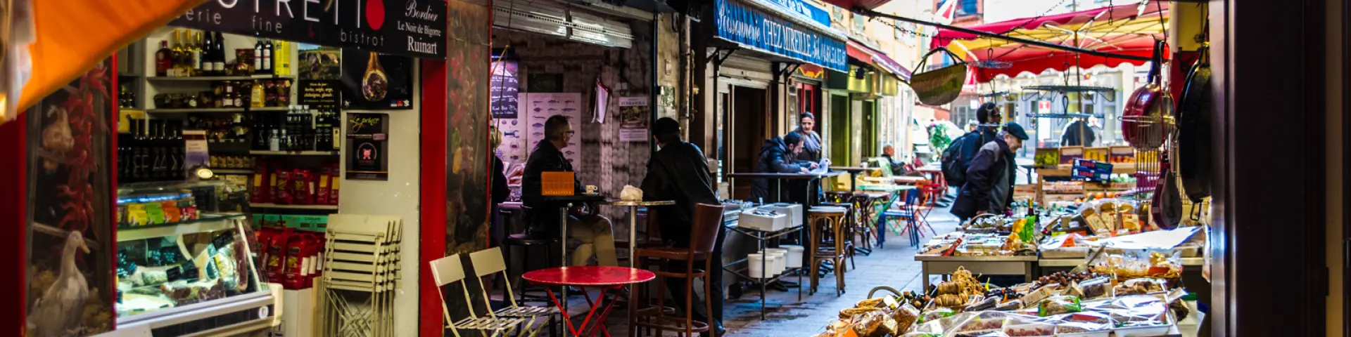 Ruelle animée bordée d’étals et de terrasses colorées où des passants discutent et dégustent des produits locaux.