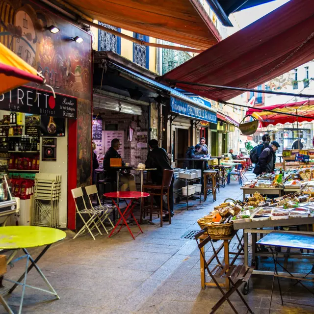 Ruelle animée bordée d’étals et de terrasses colorées où des passants discutent et dégustent des produits locaux.