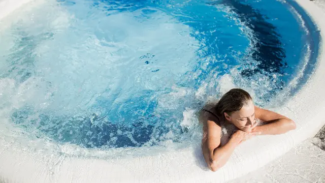 Femme se relaxant dans un bassin à remous en extérieur, appuyée sur le bord du jacuzzi, sous un soleil éclatant.