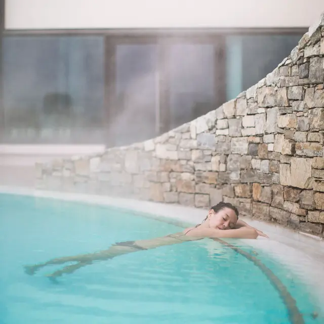 Femme se relaxant dans un bassin d’eau chaude intérieure, adossée à un mur en pierres, dans une ambiance paisible et légèrement brumeuse.