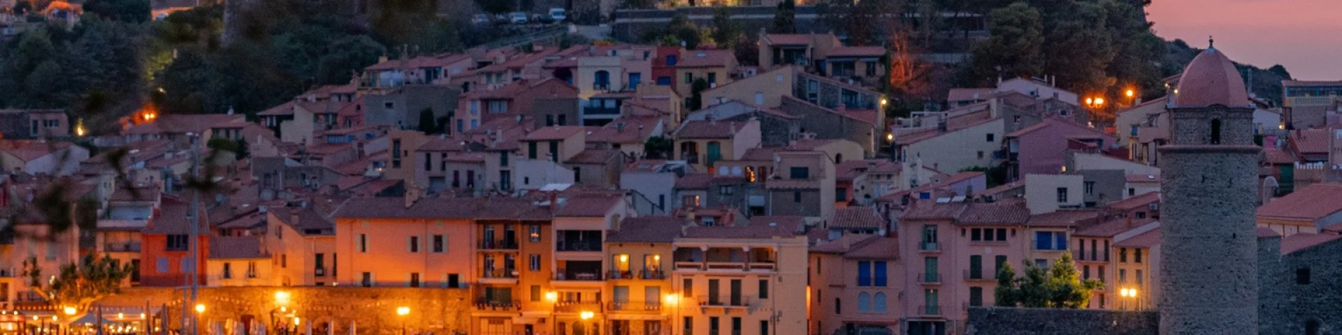 Vue sur le port et les maisons colorées de Collioure au crépuscule, avec le clocher et la mer éclairés par les lumières du soir, encadrés par des pins.