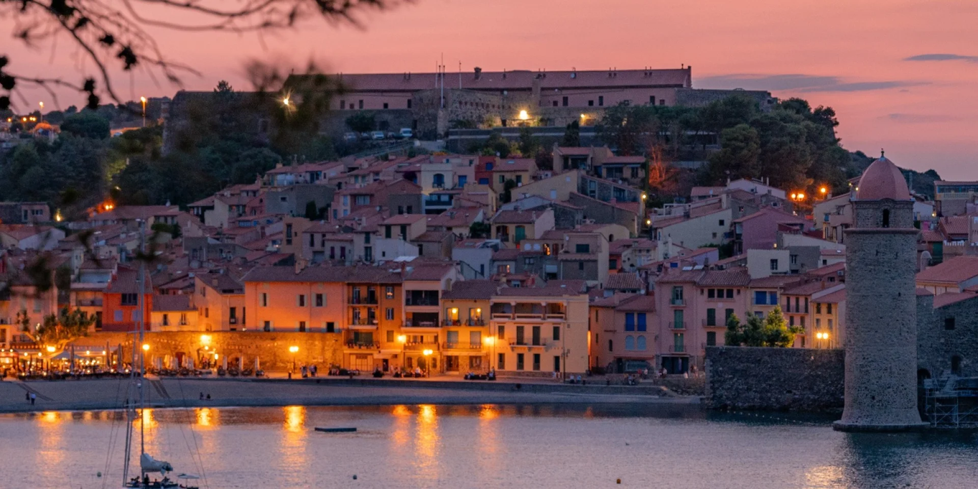 Vue sur le port et les maisons colorées de Collioure au crépuscule, avec le clocher et la mer éclairés par les lumières du soir, encadrés par des pins.