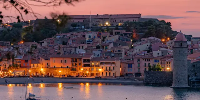 Vue sur le port et les maisons colorées de Collioure au crépuscule, avec le clocher et la mer éclairés par les lumières du soir, encadrés par des pins.