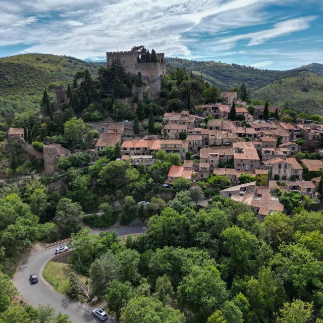 Vue aérienne du village médiéval de Castelnou dominé par son château, entouré de collines verdoyantes et de forêts sous un ciel légèrement nuageux.